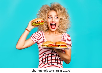 Studio Fashion Portrait Of A Beautiful Funny Young Girl On The Turquoise Background, Blonde Holding A Tray Burger Is Going To Bite A Hamburger, Curly Hair, Perfect Makeup, Crazy Emotions, Fast Food