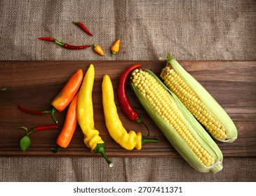 Studio flat lay of two husked corn cobs and various colorful peppers on a dark cutting board. The composition, set against a burlap background, emphasizes rustic texture and fresh ingredients, with am