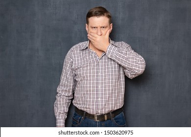Studio half-length portrait of angry man wearing checkered shirt, covering mouth with hand like not wanting to say something, over gray background. Concept of deliberate silence and reticence - Shutterstock ID 1503620975