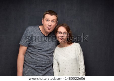 Studio half-length portrait of funny fair-haired caucasian couple grimacing and showing tongues while joking, teasing and having fun together, looking cheerfully at camera over gray background