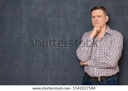 Studio half-length portrait of serious focused man wearing shirt, propping head with fist, looking thoughtfully at camera, standing in pondering position over gray background, copy space on left