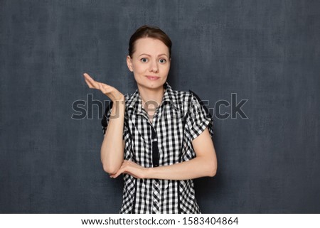 Studio half-length portrait of upset bewildered caucasian girl wearing checkered shirt, looking with puzzled expression at camera, raising hand, not understanding what happened, over gray background