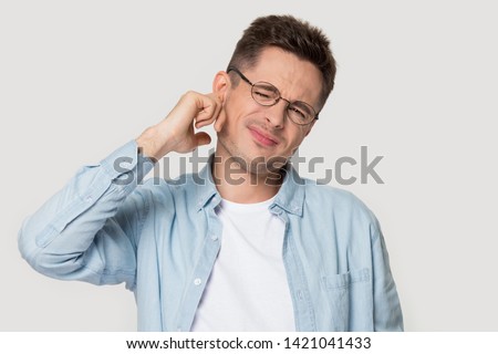 Studio head shot portrait on grey background frowning young man in glasses touch ear closed it with finger hurt suffers from earache strong pain, guy look at camera annoyed by loud noise concept image