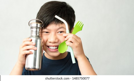 Studio and headshot : Smart looking preteen / teenage Asian boy smile and hold stainless water bottle, reusable green spoon / fork and blue silicone straw close to face. Eco-friendly Lifestyle Living.