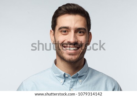 Studio headshot of young happy European Caucasian man pictured isolated on gray background with short dark hair and formal light blue shirt, laughing positively and friendly showing pleasure