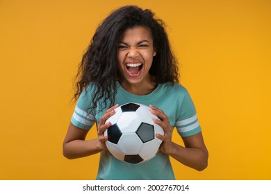Studio image of euphoric soccer fan girl holding ball in hands looking at the camera and screaming, supporting favorite team. Isolated over vivid yellow background.