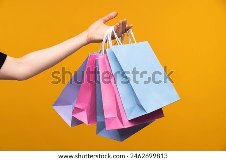 Studio image of female hand holding a bunch of pastel colored papaer shopping bags isolated over bright colored orange yellow background.