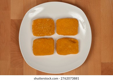 A studio image of four uncooked, breaded Trappist cheese pieces arranged on a white plate on a wooden table