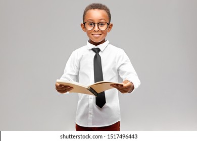 Studio Image Of Funny Smart Akward African Schoolboy In Formal Clothes And Eyeglasses Eager To Learn, Holding Open Book In His Hands And Smiling. Enthusiastic Dark Skinned A-student Learning At School