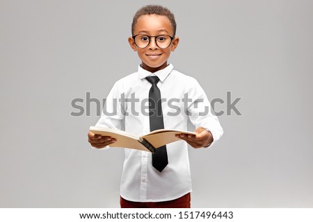 Studio image of funny smart akward African schoolboy in formal clothes and eyeglasses eager to learn, holding open book in his hands and smiling. Enthusiastic dark skinned A-student learning at school