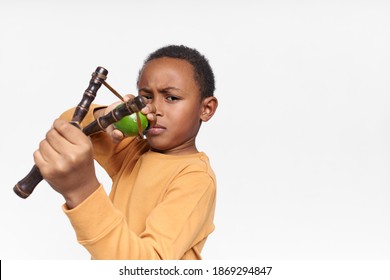 Studio image of mischievous naughty African American boy frowning having concentrated look, firing green lime from hand crafted wooden slingshot. Children, active lifestyle and games concept