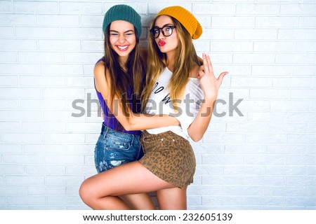 Studio lifestyle portrait of two best friends hipster girls wearing stylish bright outfits, hats, denim shorts and glasses, going crazy and having great time together. White urban wall background. 