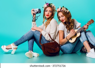 Studio lifestyle portrait of  two pretty young blonde   happy girls having fun. Making pictures in hipster style  on retro camera, wearing stylish vintage boho outfits and  flowers wreath. Spring.
