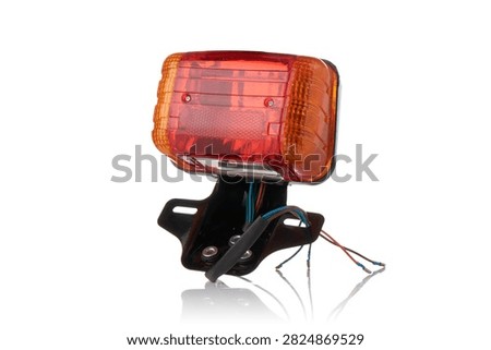 Studio macro close up of motorcycle rear tail light assembly featuring red brake light diffuser lens, amber turn signal indicators, integrated reflector, black metal license plate bracket, and wiring harness, isolated on pure white background with soft mirror reflection.