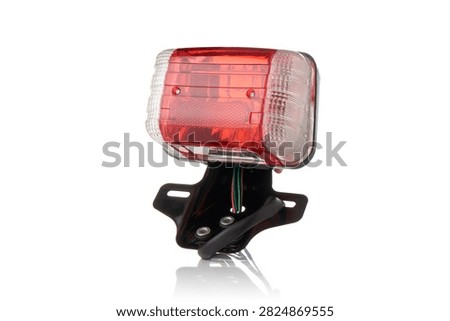 Studio macro close up of motorcycle rear tail light assembly featuring red brake light diffuser lens, amber turn signal indicators, integrated reflector, black metal license plate bracket, and wiring harness, isolated on pure white background with soft mirror reflection.