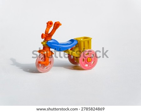 Studio macro shot of a small plastic toy tricycle, featuring an orange frame, blue seat, yellow rear basket, and pink wheels casting a soft shadow on a clean white background.