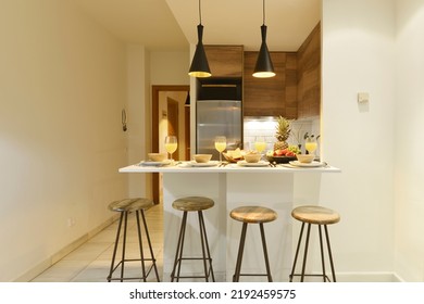 Studio With Open Kitchen, White Breakfast Bar With Juices, Industrial Wooden And Metal Stools And Fruits In Vacation Rental House