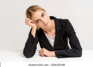 Studio Photo Of A Beautiful Blonde Girl Talking In A Business Suit On A White Background Sitting At The Table. She Is Sitting Right In Front Of The Camera, Smiling And Looking Happy.