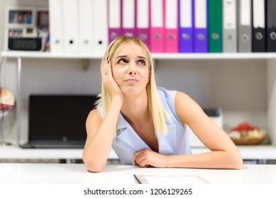 Studio Photo Of A Beautiful Blonde Girl Is Talking In A Business Suit In An Office Sitting At A Table. She Is Sitting Right In Front Of The Camera, Smiling And Looking Happy.