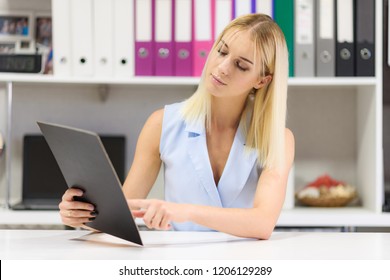 Studio Photo Of A Beautiful Blonde Girl Is Talking In A Business Suit In An Office Sitting At A Table. She Is Sitting Right In Front Of The Camera, Smiling And Looking Happy.