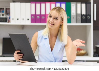 Studio Photo Of A Beautiful Blonde Girl Is Talking In A Business Suit In An Office Sitting At A Table. She Is Sitting Right In Front Of The Camera, Smiling And Looking Happy.