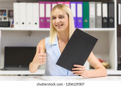 Studio Photo Of A Beautiful Blonde Girl Is Talking In A Business Suit In An Office Sitting At A Table. She Is Sitting Right In Front Of The Camera, Smiling And Looking Happy.