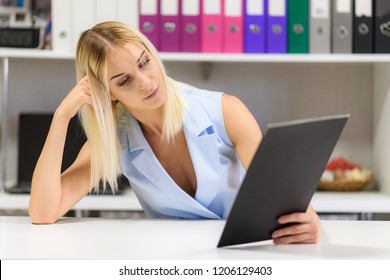 Studio Photo Of A Beautiful Blonde Girl Is Talking In A Business Suit In An Office Sitting At A Table. She Is Sitting Right In Front Of The Camera, Smiling And Looking Happy.