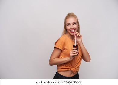 Studio Photo Of Cheerful Young Attractive Woman In Orange T-shirt Drinking Soda With Straw While Looking Aside With Charming Smile, Posing Against White Background