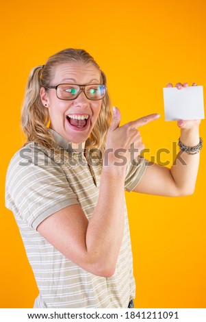 Studio photo of geeky blonde girl with glasses pointing to a blank card on yellow background.