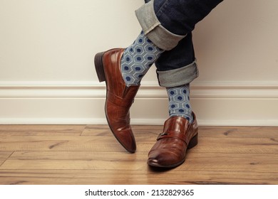 Studio photo of men's legs wearing jeans, colourful socks and brown shoes. His jeans are rolled up at the bottom. The background is grey. 