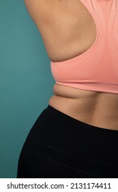Studio photo of a woman's body wearing a pink sports bra and black yoga pants. There is rolls on her waistline. The woman is caucasian. 