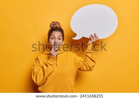 Studio photo of young thoughtful beautiful fit European woman standing in centre isolated on yellow background holding white speech bubble with blank space for your advertisement wearing hoodie