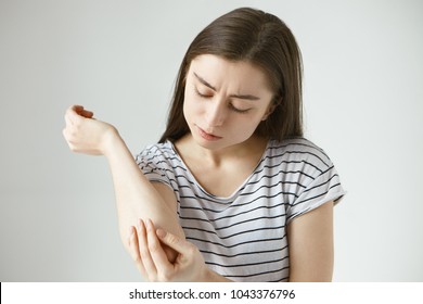 Studio Picture Of Frustrated Young Dark Haired Female Studying Skin On Her Arm After She Fell Off Bike. Student Girl Dressed In Stylish Striped T-shirt Looking At Her Elbow, Feeling Itch Or Pain