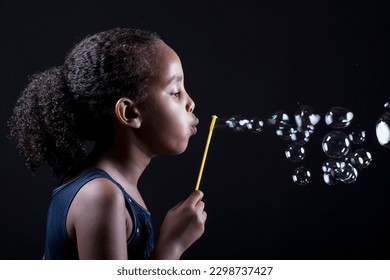  A studio portrait of a 10-year-old African American girl of Ethiopian descent blowing bubbles, with her curly hair in a ponytail and a black background adding contrast.