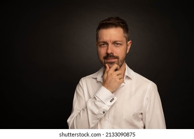 Studio Portrait Of A 40 Year Old Caucasian Man Wearing A White Shirt. The Background Is Black. He Is Looking At The Camera And Has His Hand Touching His Chin. His Expression Looks Like He Is Thinking.