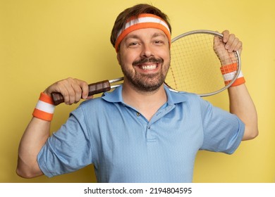 Studio Portrait Of A 40 Year Old Attractive Caucasian Man Wearing Vintage Clothing And Holding A Vintage Tennis Racket. The Studio Background Is Yellow.