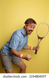 Studio Portrait Of A 40 Year Old Attractive Caucasian Man Wearing Vintage Clothing And Holding A Vintage Tennis Racket. The Studio Background Is Yellow.