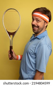 Studio Portrait Of A 40 Year Old Attractive Caucasian Man Wearing Vintage Clothing And Holding A Vintage Tennis Racket. The Studio Background Is Yellow.