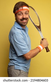 Studio Portrait Of A 40 Year Old Attractive Caucasian Man Wearing Vintage Clothing And Holding A Vintage Tennis Racket. The Studio Background Is Yellow.