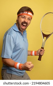 Studio Portrait Of A 40 Year Old Attractive Caucasian Man Wearing Vintage Clothing And Holding A Vintage Tennis Racket. The Studio Background Is Yellow.