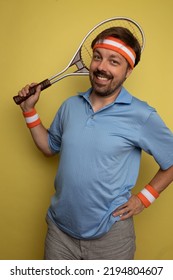 Studio Portrait Of A 40 Year Old Attractive Caucasian Man Wearing Vintage Clothing And Holding A Vintage Tennis Racket. The Studio Background Is Yellow.