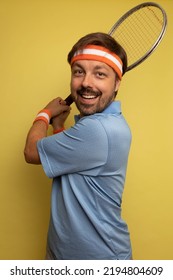 Studio Portrait Of A 40 Year Old Attractive Caucasian Man Wearing Vintage Clothing And Holding A Vintage Tennis Racket. The Studio Background Is Yellow.