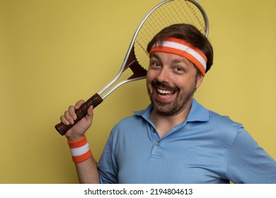 Studio Portrait Of A 40 Year Old Attractive Caucasian Man Wearing Vintage Clothing And Holding A Vintage Tennis Racket. The Studio Background Is Yellow.