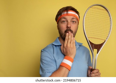 Studio Portrait Of A 40 Year Old Attractive Caucasian Man Wearing Vintage Clothing And Holding A Vintage Tennis Racket. The Studio Background Is Yellow.