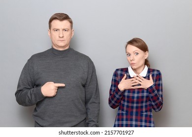 Studio Portrait Of Adult Couple, Serious Mature Man Is Pointing With Index Finger At Surprised Woman Looking Confused And Unawared, With Widened Eyes And Eyebrows Raised. Relationship Concept