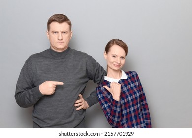 Studio Portrait Of Adult Couple, Serious Mature Man Is Pointing With Index Finger At Happy Woman Pointing With Thumb At Man And Smiling Cheerfully, Looking Contented And Joyful. Relationship Concept