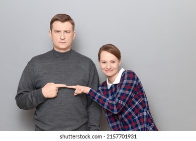 Studio Portrait Of Adult Couple, Serious Mature Man Is Pointing With Index Finger At Happy Woman Smiling Cheerfully, Looking Contented And Joyful, Having Fun And Fooling Around. Relationship Concept