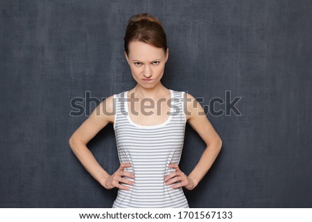 Studio portrait of angry enraged girl wearing tank top, looking disapprovingly from under forehead, holding hands on hips, being dissatisfied of her boyfriend's behavior, standing over gray background