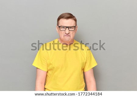Studio portrait of angry irritated blond mature man with glasses, wearing yellow T-shirt, with negative expression, grimacing from anger and rage, looking furious, standing over gray background 