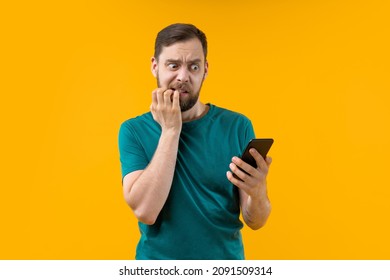 Studio portrait of anxcious shocked nervous scared man reading message on his mobile phone, eyes wide open, biting nails. Isolated over yellow background.